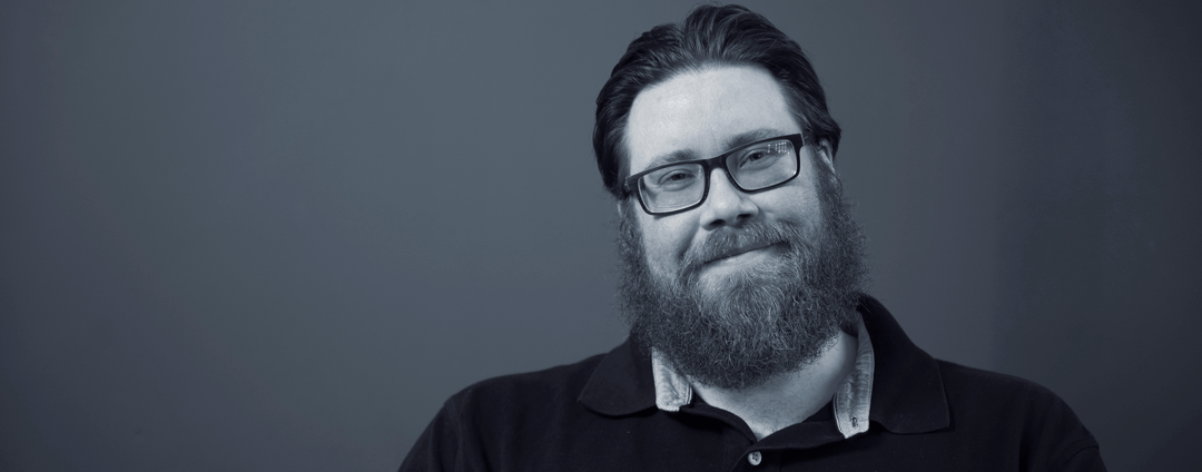 Black and white photo of a man in his thirties with a full beard and black glasses wearing a dark sweater. He is smiling at the camera