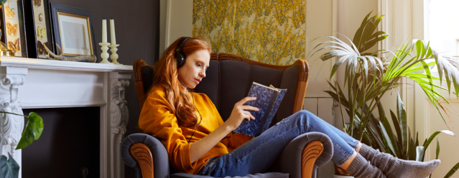Young red-headed woman lounging in an armchair listening to headphones and reading a book