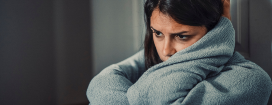 A distressed young adult female in a gray sweater, sitting against a door frame, holding her knees to her chest and resting her head on her left arm.