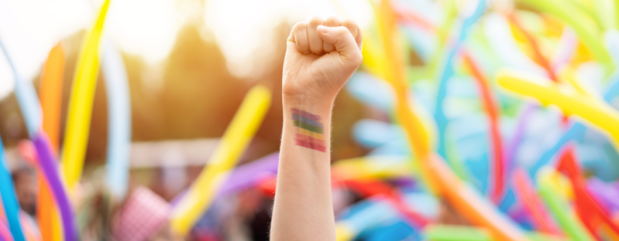 A white person's arm in the air with a rainbow tattoo on their wrist. Looks like they are attending a festival.