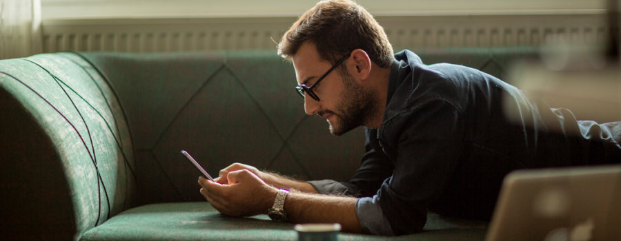 A white male with light brown hair, beard, and glasses lays on his stomach on a green couch reading on his cell phone.