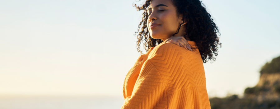 Young woman in a golden yellow sweater standing on a beach with her eyes closed and holding her right hand over her heart. Her curly hair is tussled in the wind.