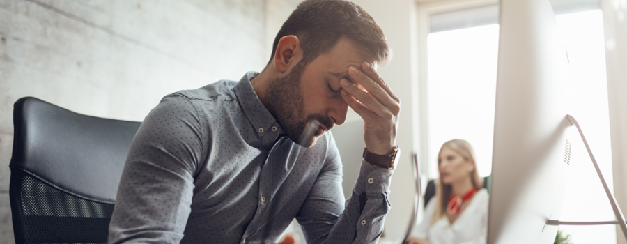 Picture of a man sitting at his desk, holding his head out of frustration with his co-worker sitting in the background.