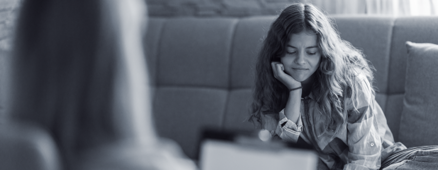 Black and white photo of pre-teen girl with curly light brown hair, sitting on a couch with her chin in her palm, slightly smiling. A grown up adult woman is sitting across from her in a chair holding a clipboard.