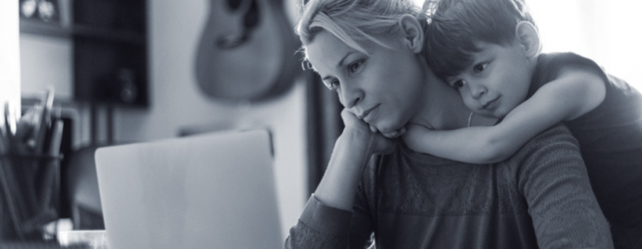 Photo of a mom sitting at her laptop computer looking worried. Her young son has his arms wrapped around her neck with a soft smile on his face.