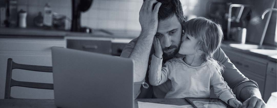 Photo of a dad sitting at his laptop computer looking stressed out. His head is in his hand and his daughter is on his lap kissing his cheek.