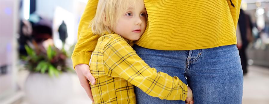 Young girl with blond hair and blue eyes wearing a yellow plaid sweater is holding onto parents leg, expressing anxiety. Parent is wearing yellow sweater and blue jeans,