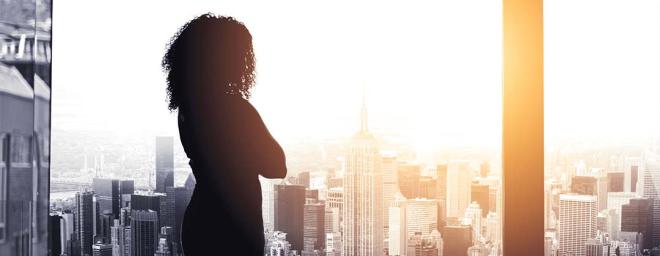 woman standing in an office building looking out a large picture window at city skyscrapers