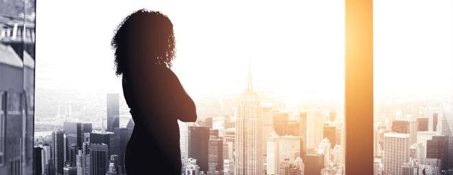 woman standing in an office building looking out a large picture window at city skyscrapers