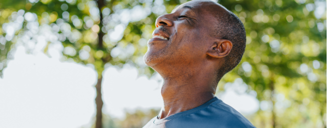 An adult Black male deeply breathing in fresh air while outside under a tree