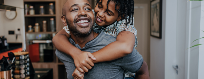 Black young boy hanging on the back of his father. Both father and son are smiling.