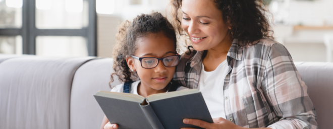 An adult woman holding open a book with a child sitting in front of the adult reading the book.