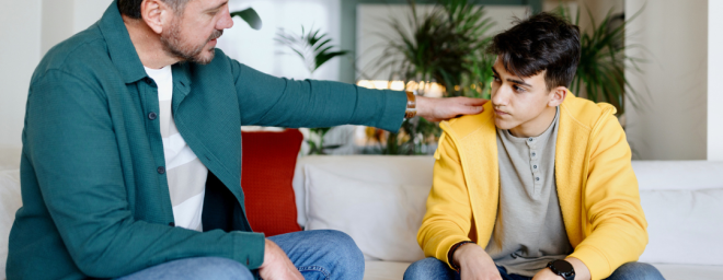 A dad holding the shoulder of a male teenager while sitting on a couch.