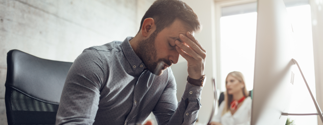 Picture of a man sitting at his desk, holding his head out of frustration with his co-worker sitting in the background.