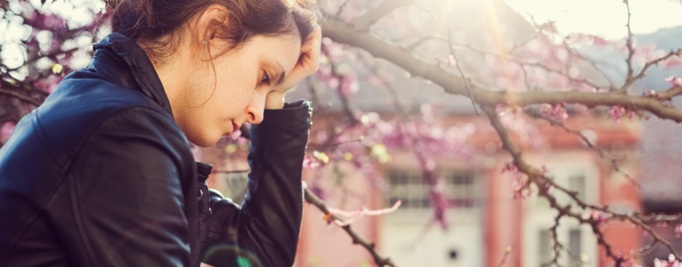 Young woman sitting outside next to a tree with her head in her hand, looking depressed.
