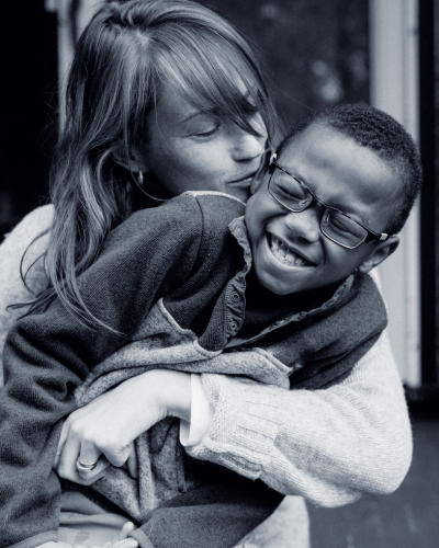 Black and white photos of a white female with light brown hair hugging a Black boy and kissing his cheek. Black boy looks to be about seven years old and is wearing black plastic glasses