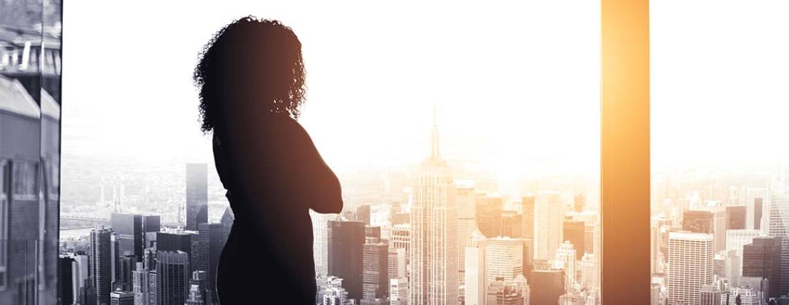 woman standing in an office building looking out a large picture window at city skyscrapers