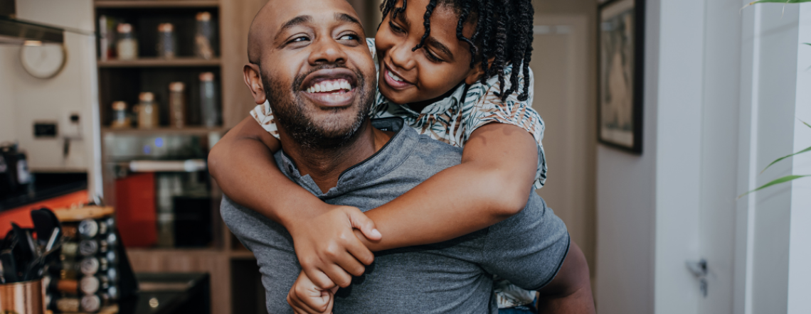 Black young boy hanging on the back of his father. Both father and son are smiling.