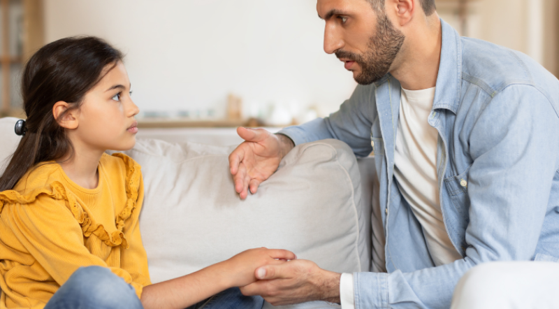 An adult male/dad talking to you younger female child while holding her hand