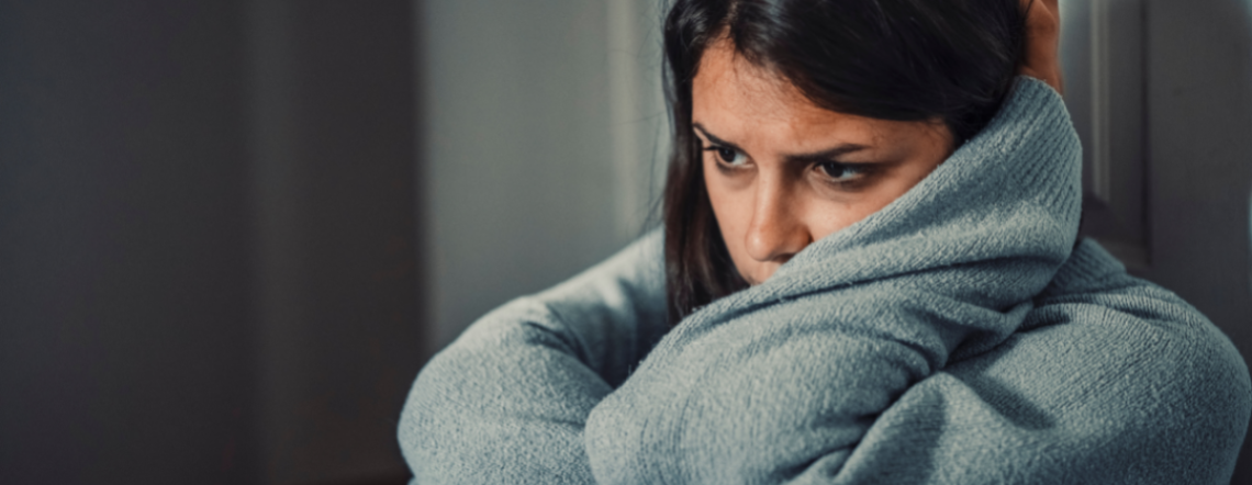A distressed young adult female in a gray sweater, sitting against a door frame, holding her knees to her chest and resting her head on her left arm.